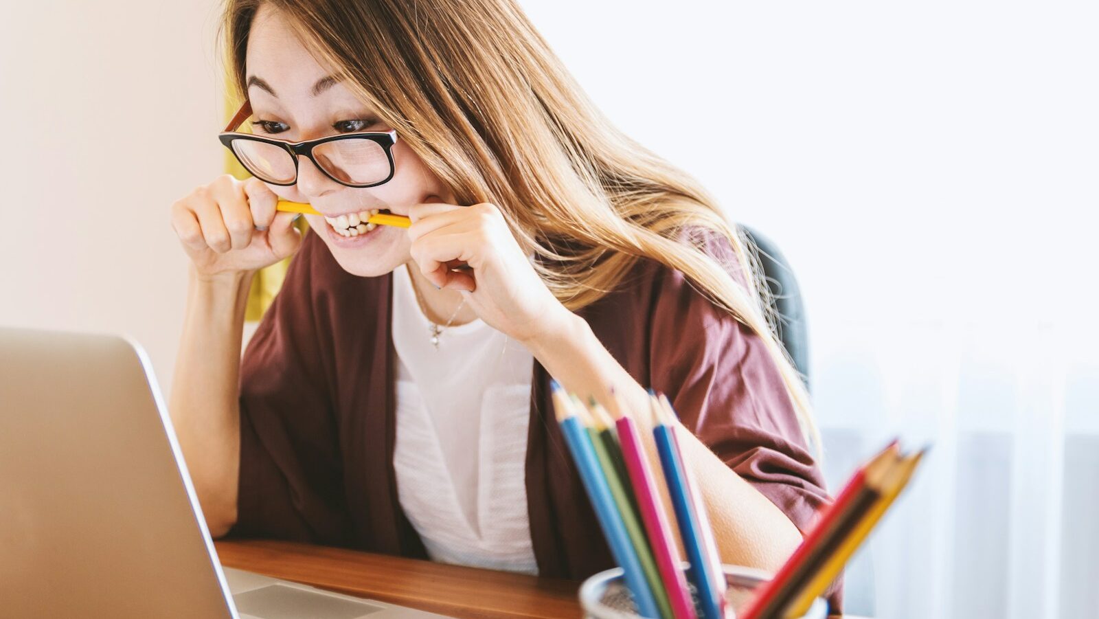 woman biting pencil while sitting on chair in front of computer during daytime